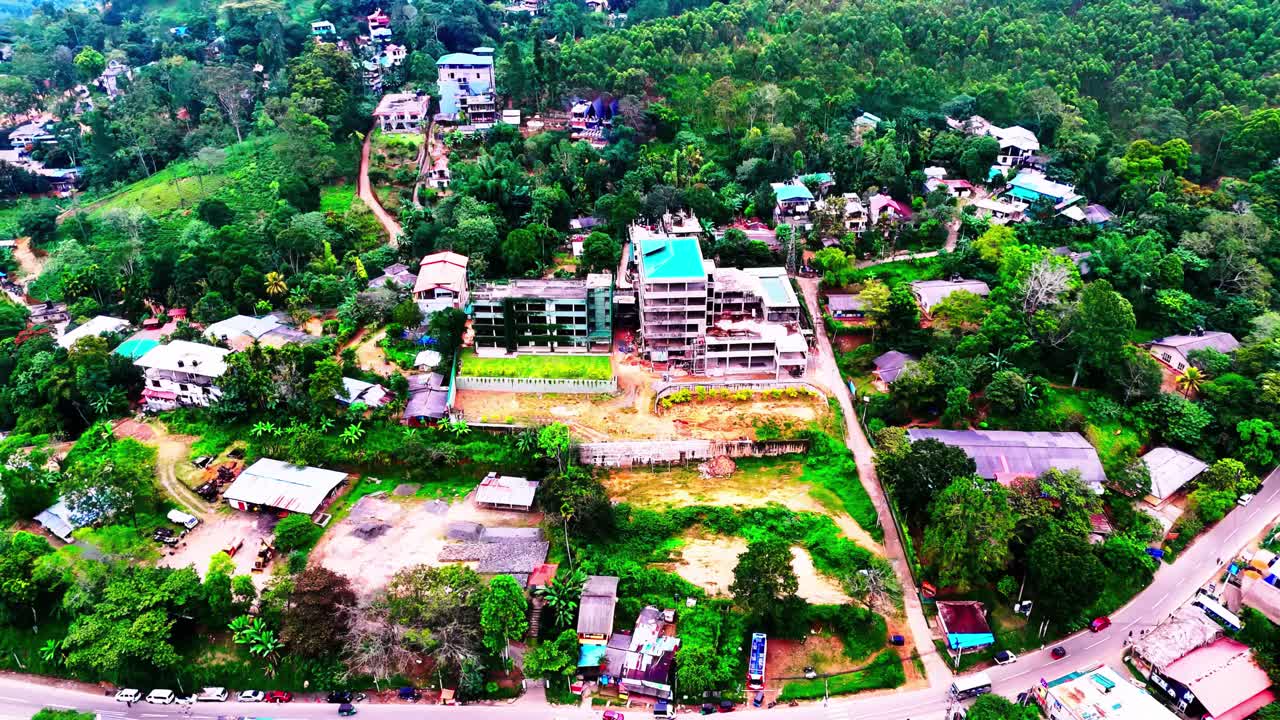A residential complex under construction emerges from forested terrain near a Sri Lankan town, surrounded by winding roads, tropical trees, and scattered village homes.