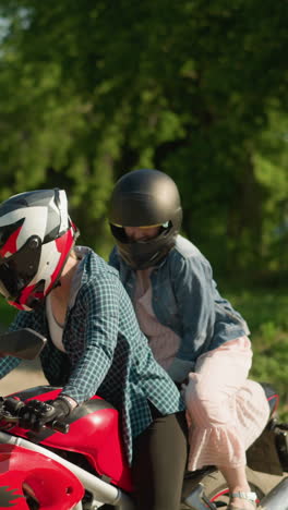 a woman in a checkered shirt and helmet sits on a red power bike with her friend, who is wearing a pink gown and blue jacket, on a quiet, tree-lined paved road