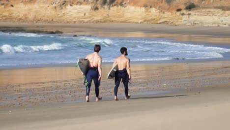 handheld shot of two male sporty surfers with their surfboards running along a charming sandy beach while calm waves in blue sea