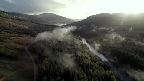 Drohnenaufnahmen-Aus-Der-Luft-Hoch-über-Den-Baumkronen-Eines-Nadelwaldes-Und-Eines-Flusses,-Der-Langsam-Hinabsteigt,-Während-Tief-Hängende-Wolken-Die-Baumwipfel-Umarmen-Und-Die-Sonne-Hinter-Bergen-Am-Horizont-Untergeht