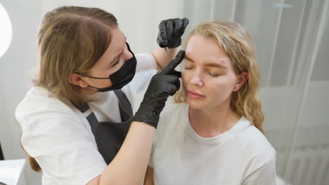 beautician in mask and gloves carefully shapes client eyebrow with tweezers, turning to work on other side. client remains calm with eyes closed, seated near soft-lit background with sheer curtain