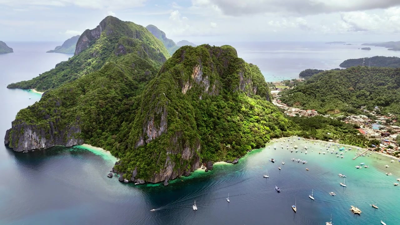 Vista aerea con drone di un grande promontorio carsico calcareo e del lungomare della città di El Nido con molte barche ancorate nella baia turchese sotto cieli nuvolosi, El Nido, Palawan, Filippine