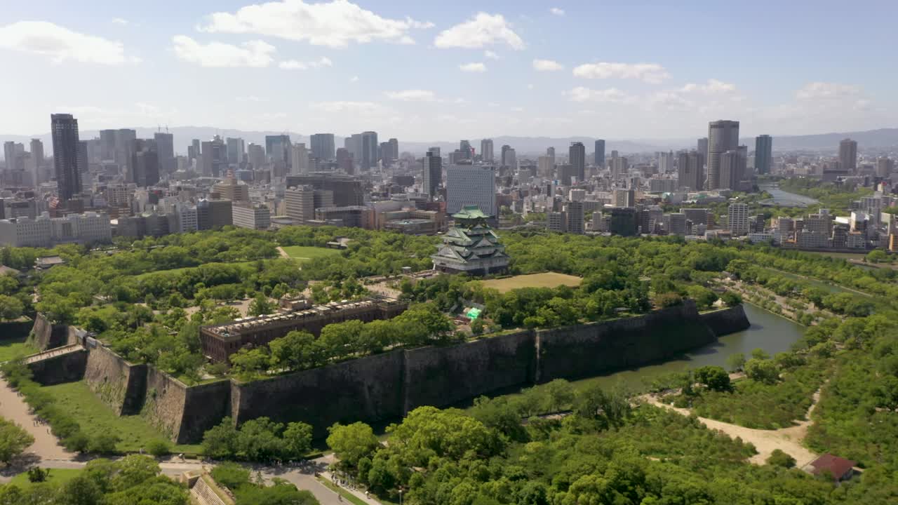 Wide erial flying towards historic Osaka Castle with park, moat, skyscraper, and urban city in Osaka, Japan