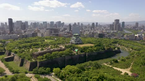 Wide-erial-flying-towards-historic-Osaka-Castle-with-park,-moat,-skyscraper,-and-urban-city-in-Osaka,-Japan