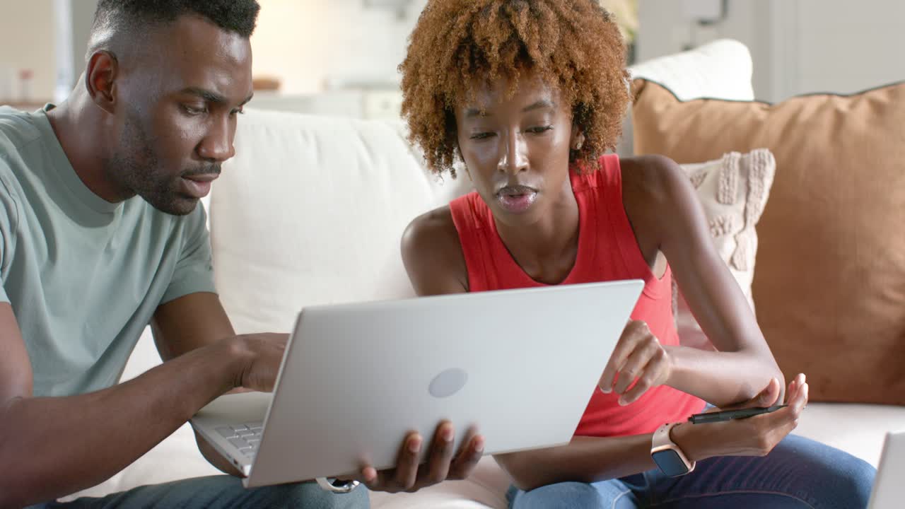 African American woman red top holding silver laptop pointing, African man on couch typing working