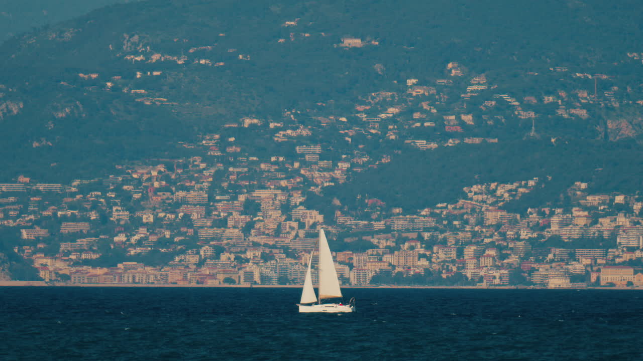 Distant view of a sail boat moving on the sea with buildings and mountains on the background