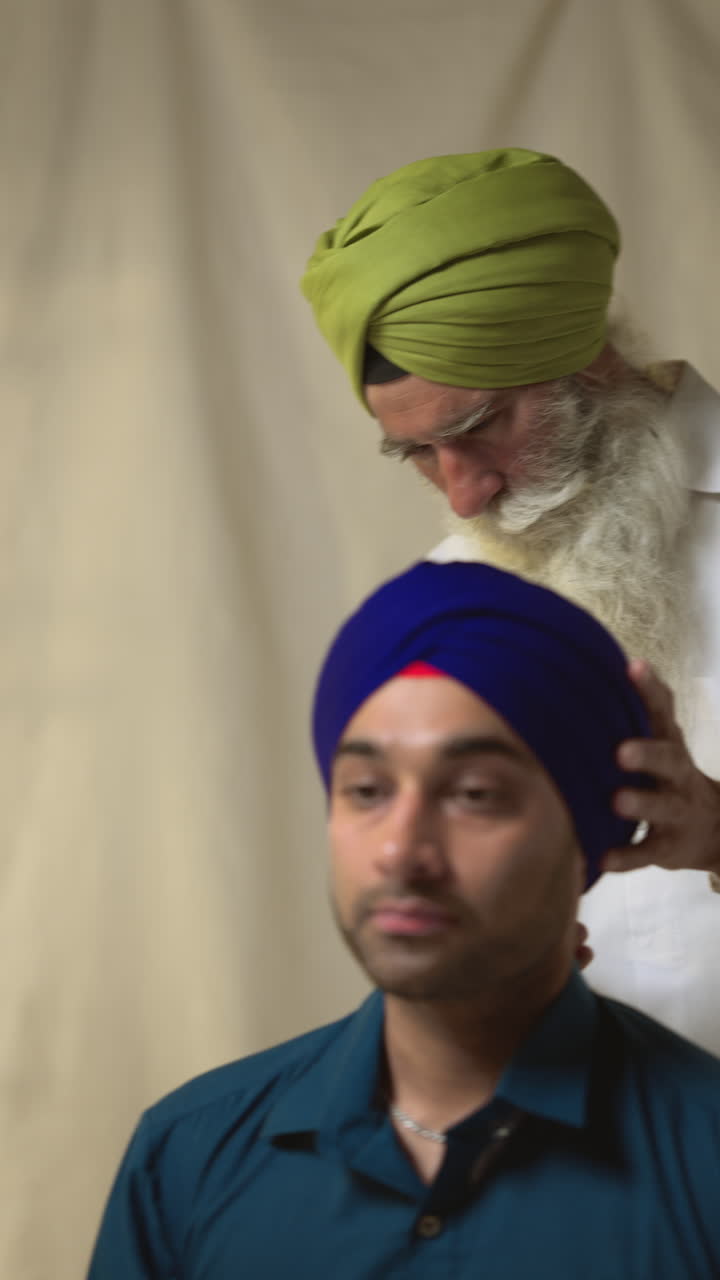 video de estudio vertical de un hombre sikh mayor usando la aguja salai en el turbante para un hombre más joven contra un fondo plano