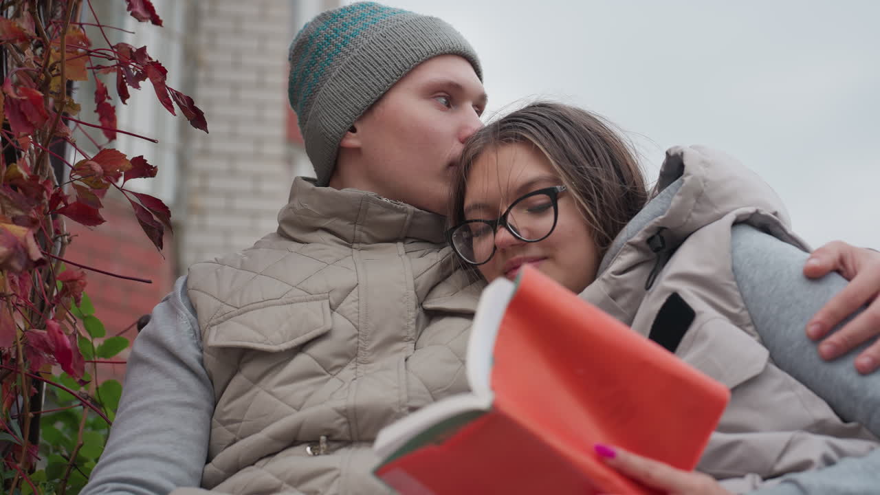Cozy moment of lovers enjoying quiet time reading together outdoors, woman resting against man chest holding open book with bright cover, surrounded by autumn leaves and greenery