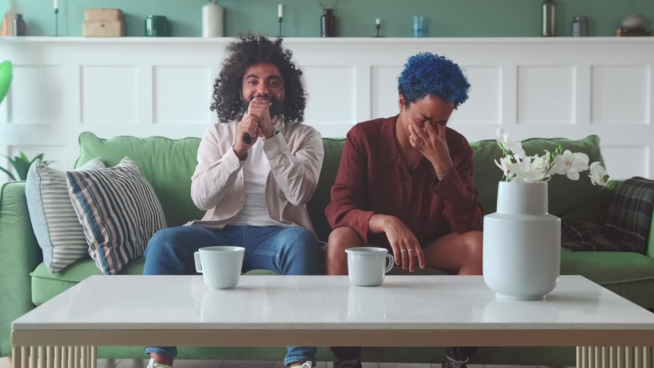 Young funny friends man and woman sing songs in karaoke sits on couch in house