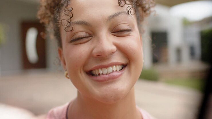 Donna afroamericana sui 20 anni che si sporge verso la fotocamera, concentrandosi, coinvolgendo e ridendo, con una canotta rosa sul portico