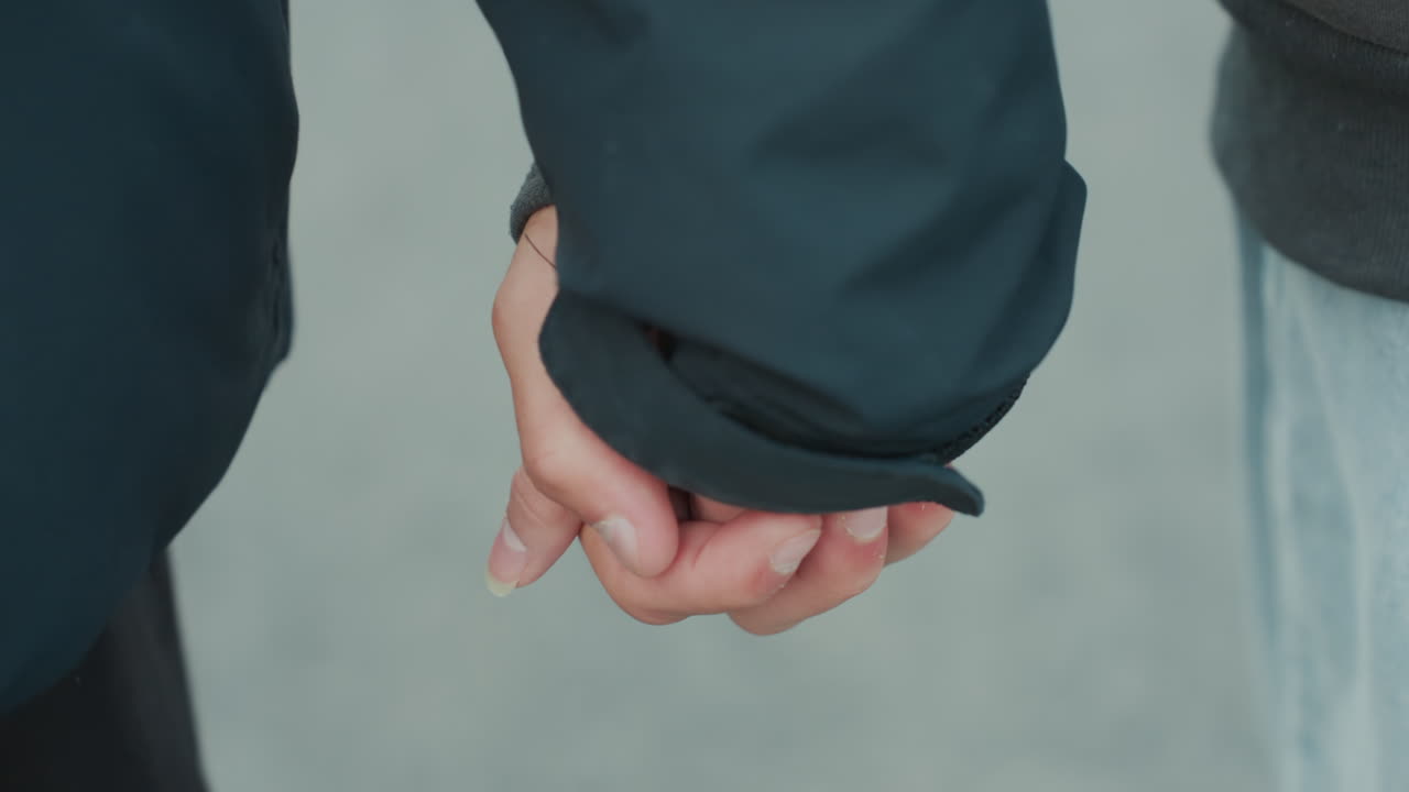 closeup hands held near waist, tight focus on fingers and palm contact against neutral pavement, dark sleeve and textured skin reveal small gestures, scene evokes commuter to confidant role change