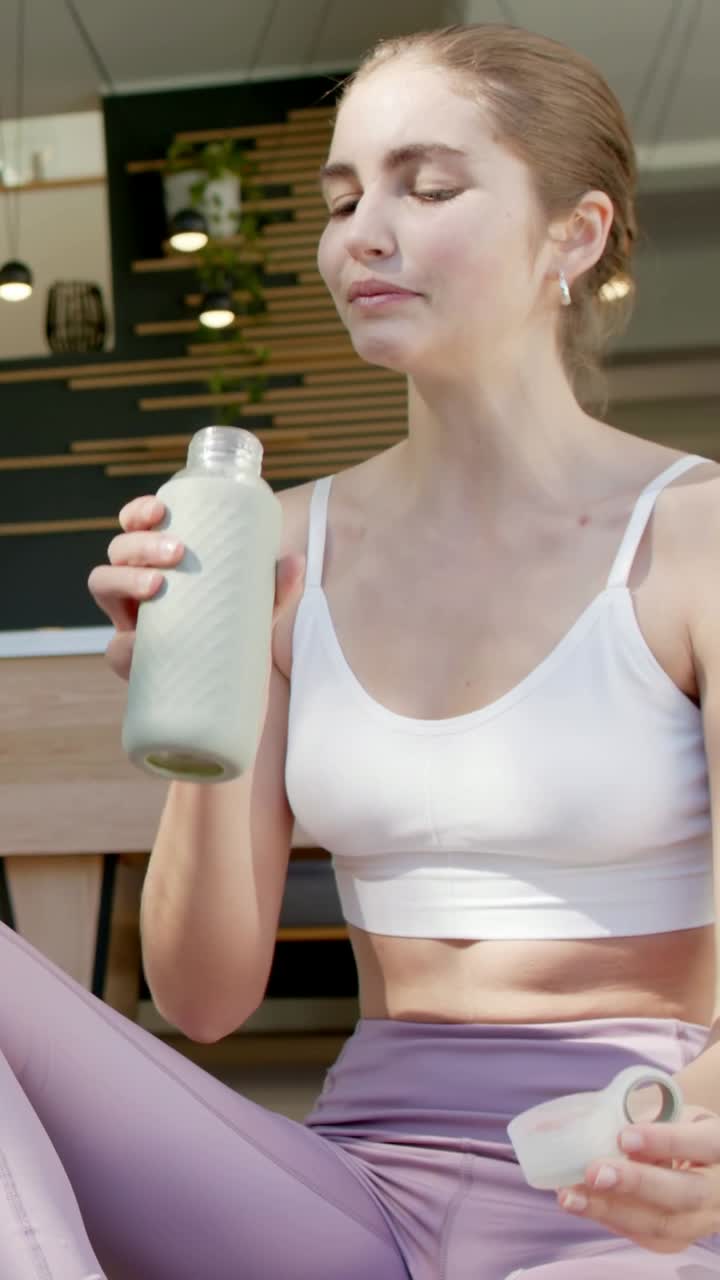 Video verticale: Donna sorridente in reggiseno e leggings lilla che avvita un tappo in un bar, sorseggiando una bottiglia per idratarsi.