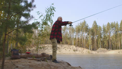 Ein-Fischer-Im-Rot-Karierten-Hemd-Wirft-Seine-Angel-Im-Flusswasser-Und-Steht-Am-Ufer-Im-Wald-Und-Angelt-An-Einem-Herbsttag-Im-Urlaub