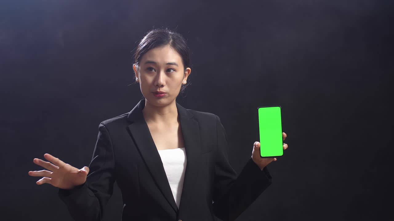 블랙 스크린 스튜디오 에서 연설 하는 동안 녹색 스크린 스마트폰 을 들고 가리키는 비즈니스 슈트 를 입은 아시아 여성