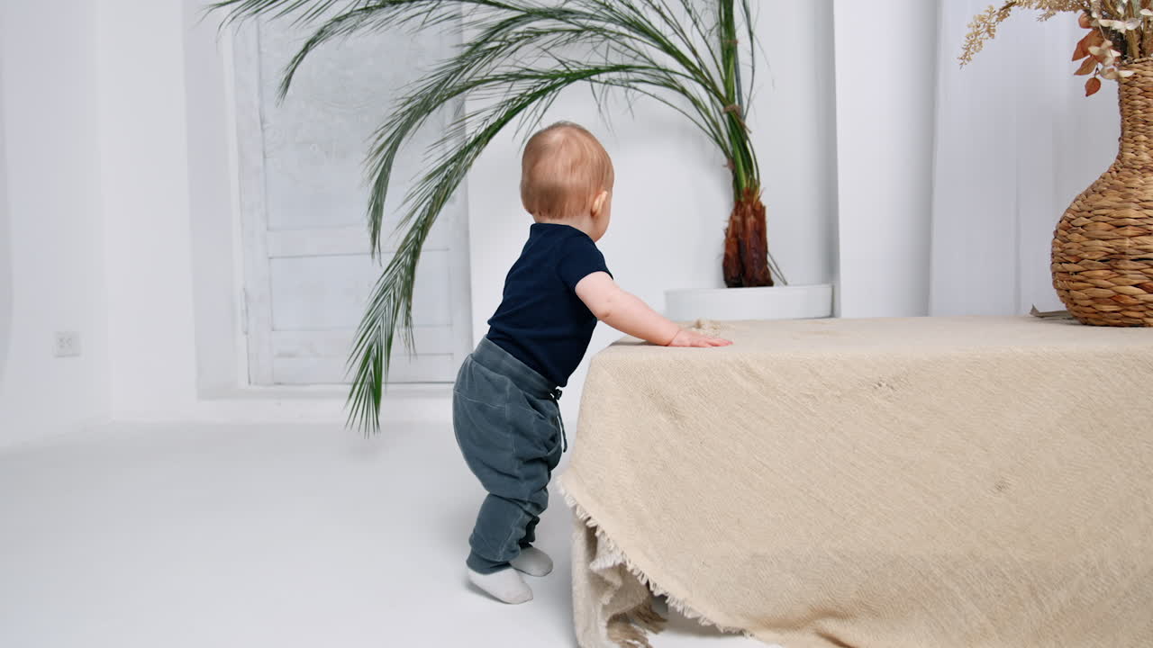 Cheerful little baby stands holding by the furniture. Infant in jeans learning to stand himself.