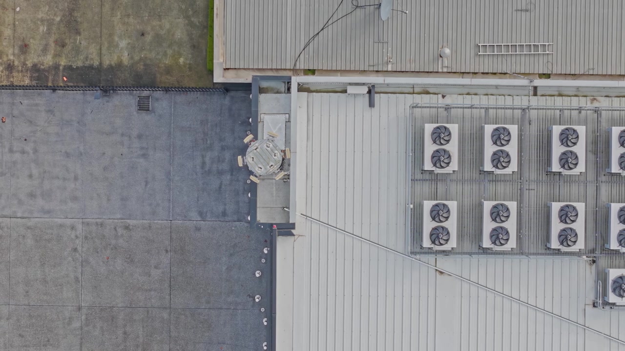 Drone video captures an overhead view of a modern data center rooftop, featuring neatly arranged HVAC cooling fans, textured concrete, industrial structures, and subtle daylight tones.
