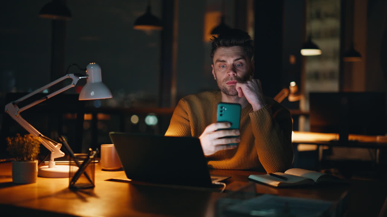 Man working on laptop and using smartphone at desk at night