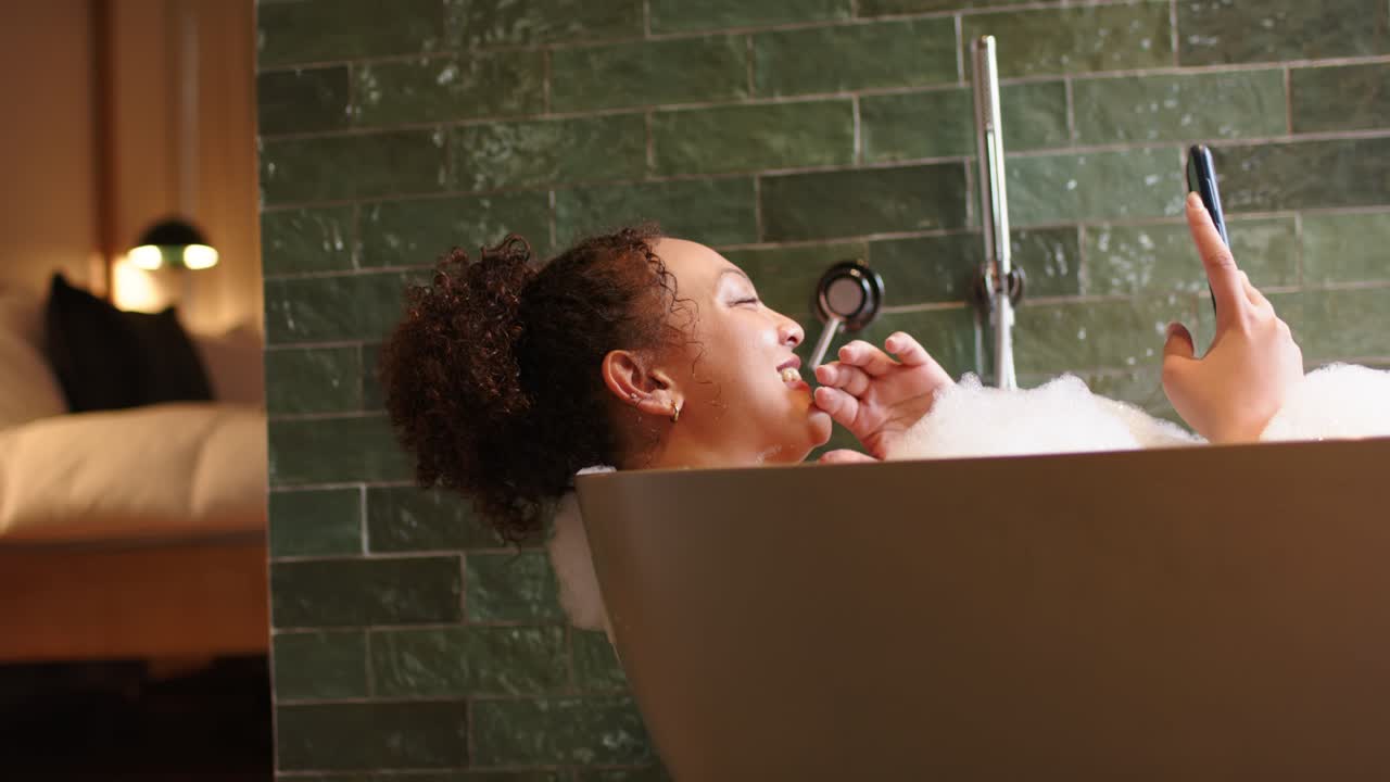 Woman is reclining in tub by green tiles, leaning back, raising phone, checking texts and laughing