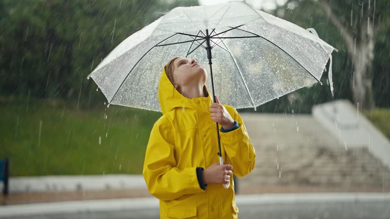 A blonde teenage girl in a yellow jacket stands under an umbrella and looks at how drops of water are dripping on him during the rain in the park