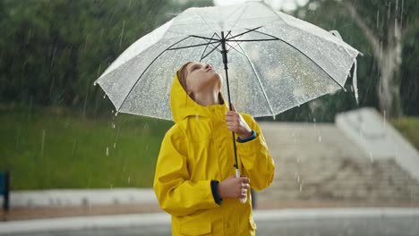 A-blonde-teenage-girl-in-a-yellow-jacket-stands-under-an-umbrella-and-looks-at-how-drops-of-water-are-dripping-on-him-during-the-rain-in-the-park