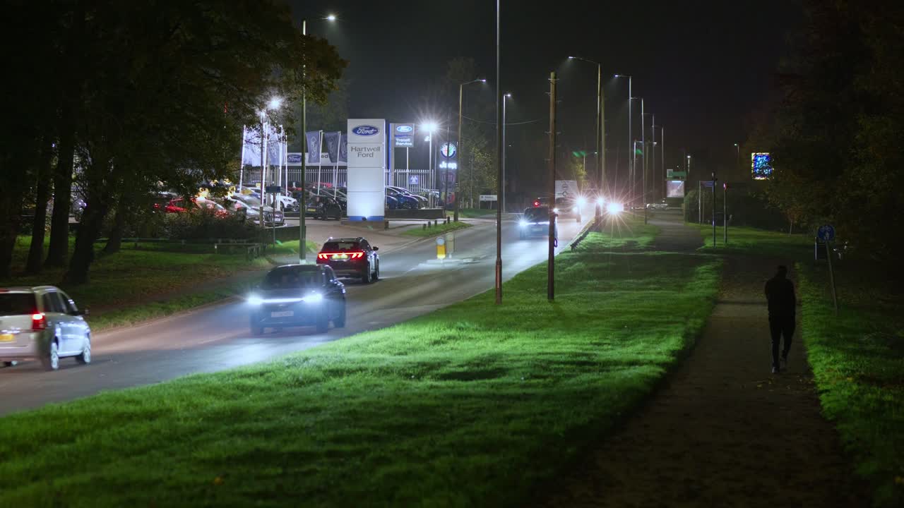 Cars with headlights drive on a night road as a pedestrian walks away on a nearby path, with a car showroom in the distance
