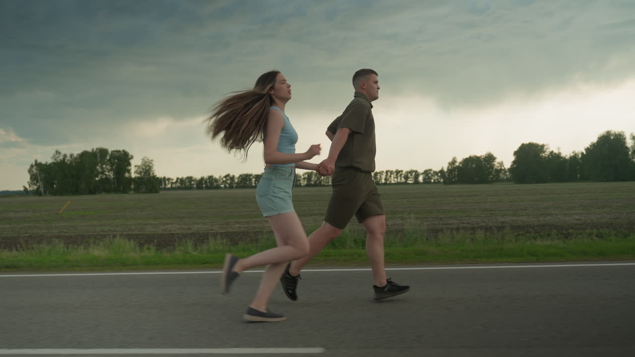 side view of man holding woman hand as he runs ahead on rural road while woman in blue tank and denim shorts chases him with hair flowing under moody cloudy sky at golden late afternoon