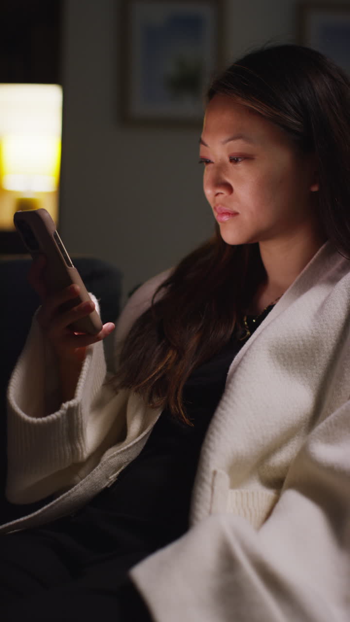 video vertical de una mujer pasando la noche en casa sentada en el sofá con el teléfono móvil desplazándose por internet o redes sociales