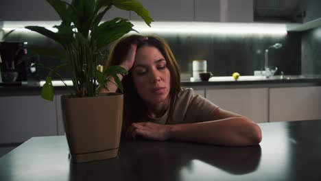 una cansada chica morena en una camiseta beige se apoya en una mesa negra y sostiene su cabeza mientras piensa cerca de una flor interior de una planta verde en la cocina en el retrato de la noche