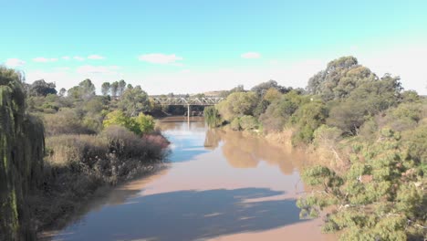 Toma-Aérea-Volando-Cerca-Del-Agua-Hacia-Un-Viejo-Puente-Oxidado-En-El-Fondo