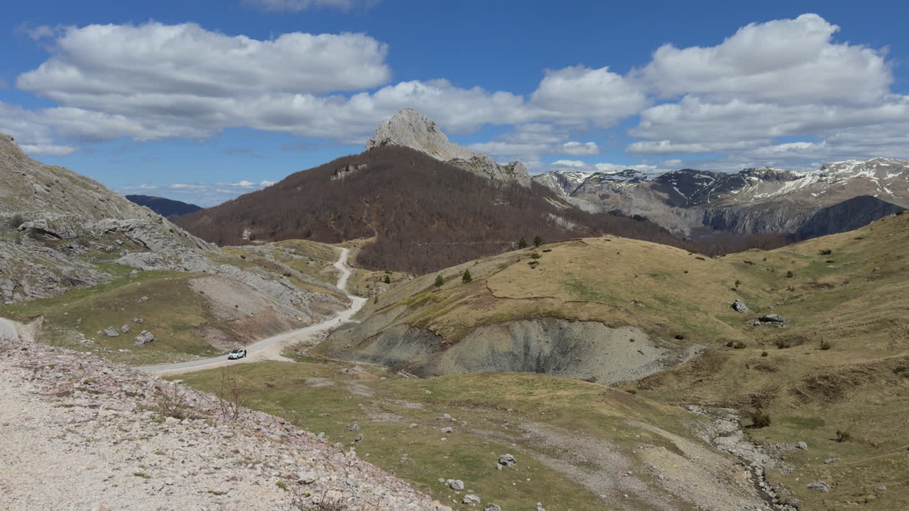A winding, narrow mountain road snakes its way through the landscape under a blue sky with clouds. Sunny day, wide shot, slow motion