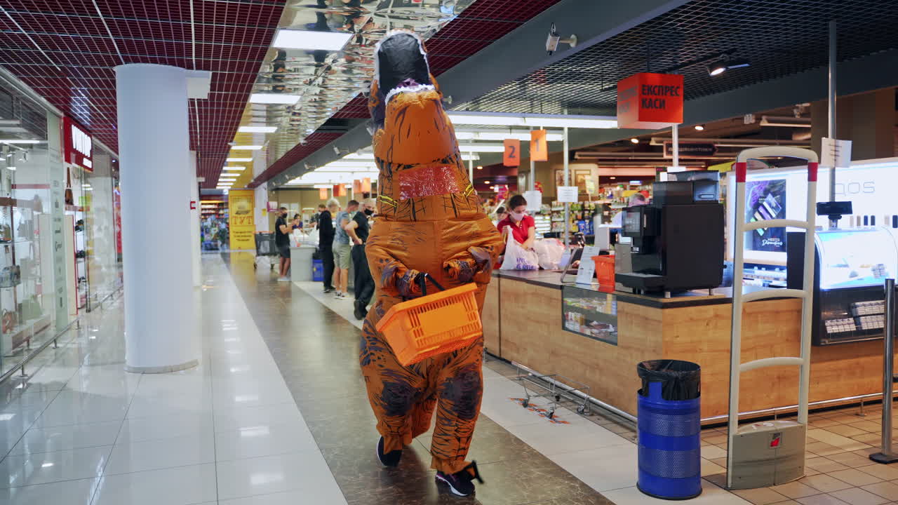 Animator in orange dinosaur costume walking by the supermarket. Man in funny inflatable costume holding a shopping basket.