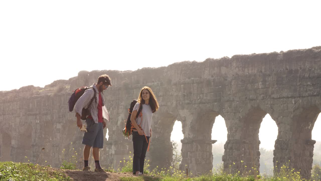 joven pareja encantadora mochileros turistas leyendo mapa señalando direcciones acueducto romano arcos en parco degli aquedotti parque ruinas en roma en romántico amanecer brumoso con guitarra y saco de dormir cámara lenta
