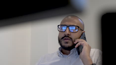 Bald-Ethnic-Minority-Entrepreneur-Talking-On-Mobile-In-Office,-Viewed-Behind-Computer-Monitors