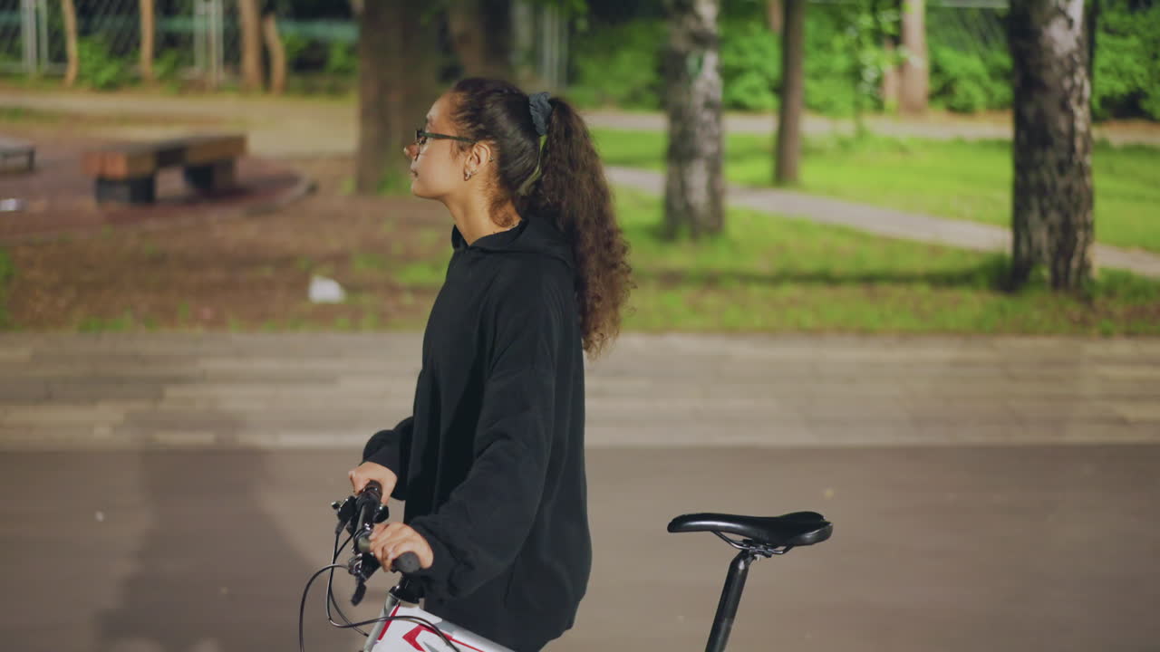 Thoughtful Female With Bicycle Outside, Contemplative Woman Holding Bike Amidst Lush Greenery And Trees, Calm Lady With Glasses Standing Beside Bicycle Surrounded By Vibrant Nature Scenes