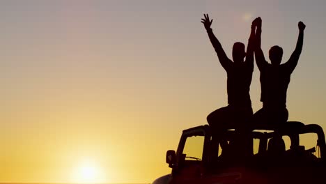 happy caucasian gay male couple sitting on car raising arms and holding hands at sunset on the beach