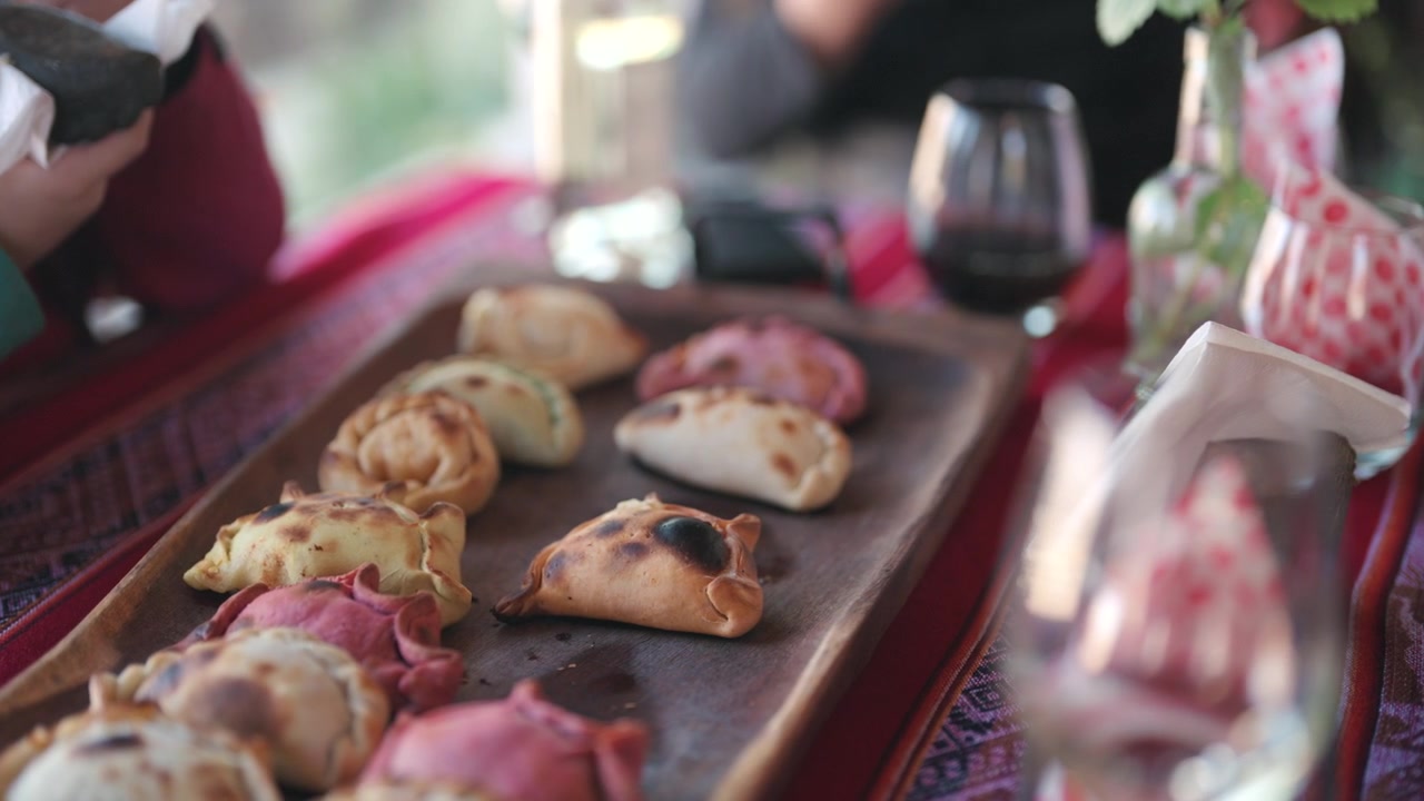 Selezione manuale di un'empanada argentina appena sfornata da un vassoio da portata rustico pieno di pasticcini assortiti durante un pasto condiviso con amici e famiglia, ripresa ravvicinata al rallentatore.