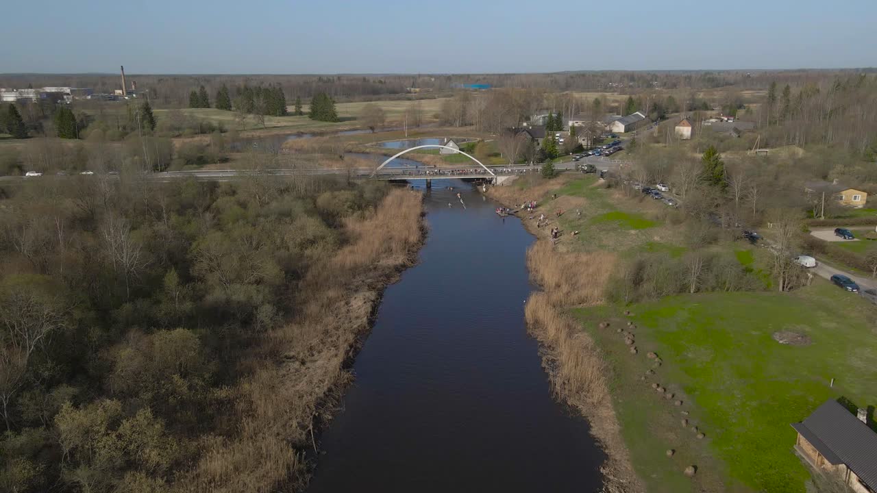 Wide overhead aerial of group kayaking on river channel next to riverside village. Forwards fly above kayakers paddle past people gathered on the shore and bridge over stream during Võhandu marathon