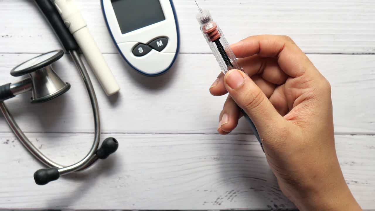 primer plano de la mano de una persona que sostiene una pluma de inyección de insulina, con un medidor de glucosa en la sangre y un estetoscopio en el fondo.