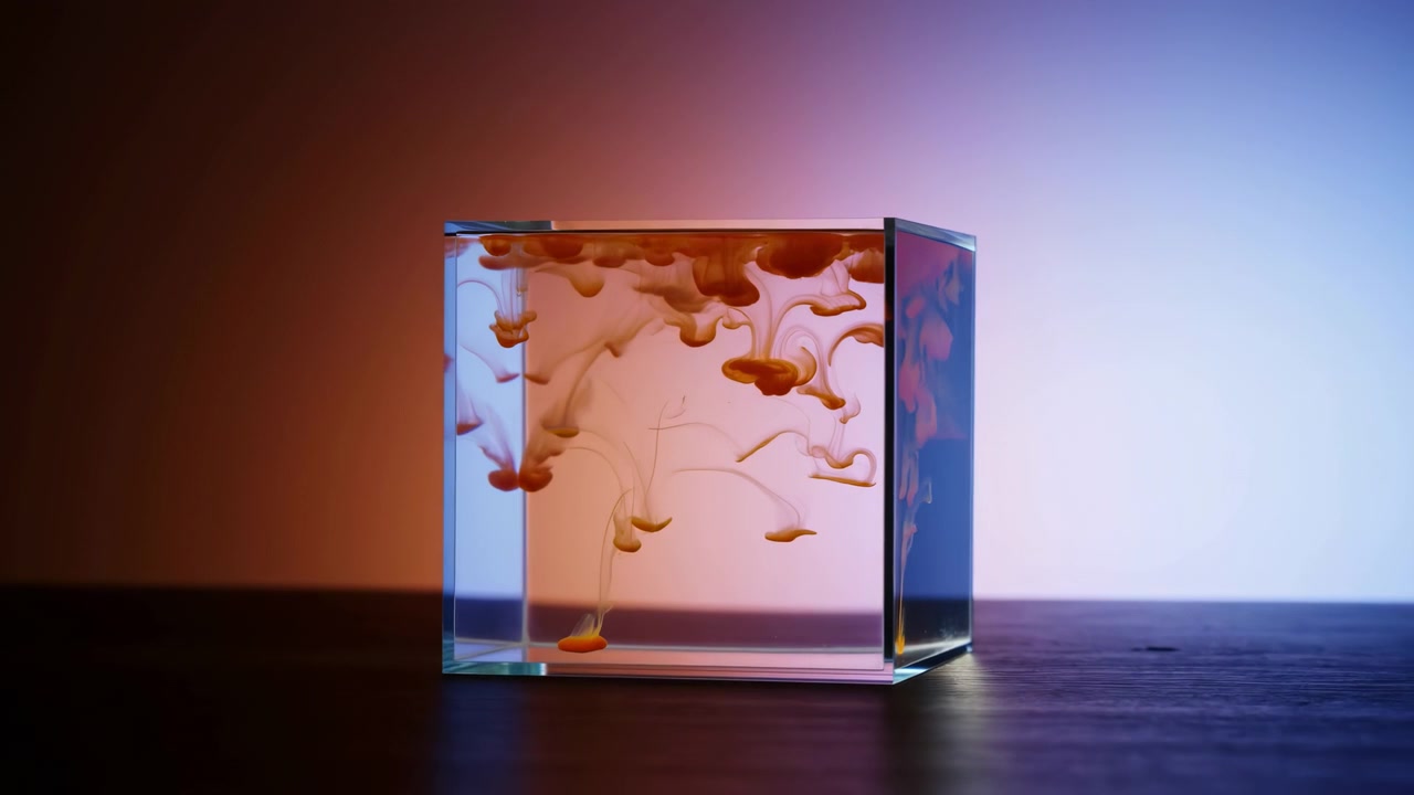 Colored fluid spreads from the upper surface into suspended wisps and rounded droplets, gradually dispersing through the clear cube container against a softly lit two-tone background.