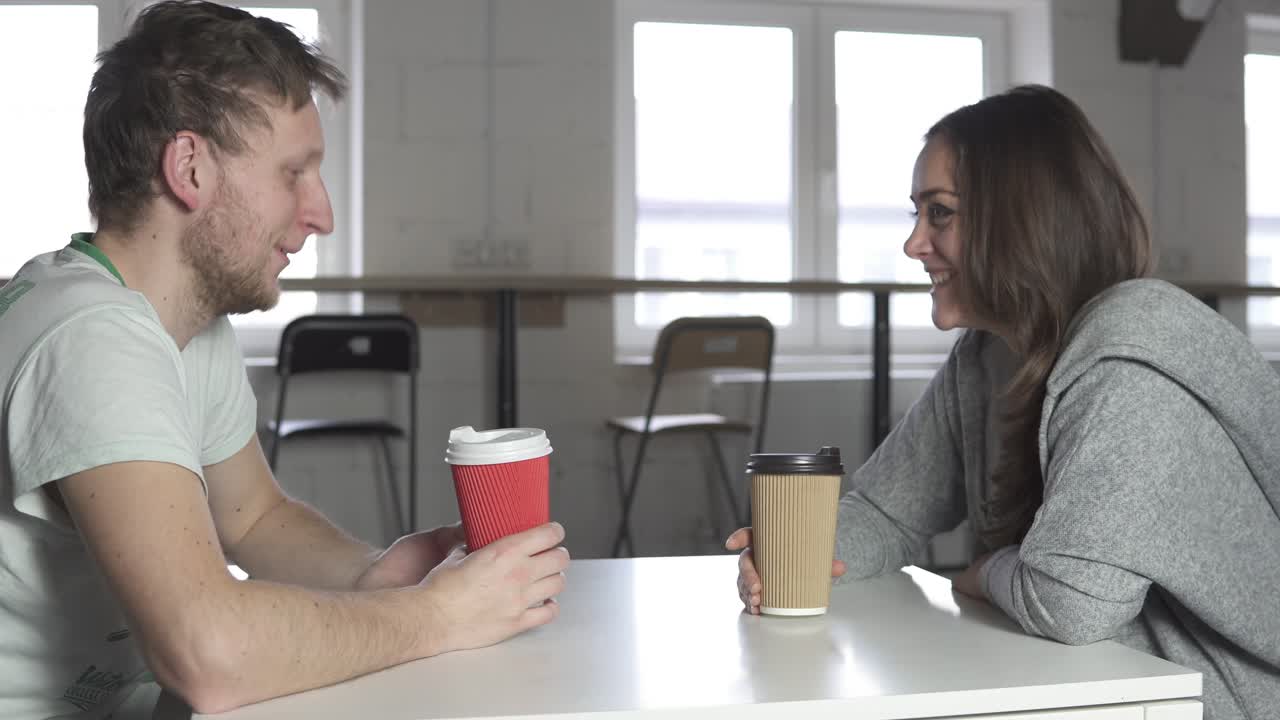 dos amigos están sentados en la mesa en una cafetería, bebiendo café para llevar en vasos de papel y hablando. filmado en 4k