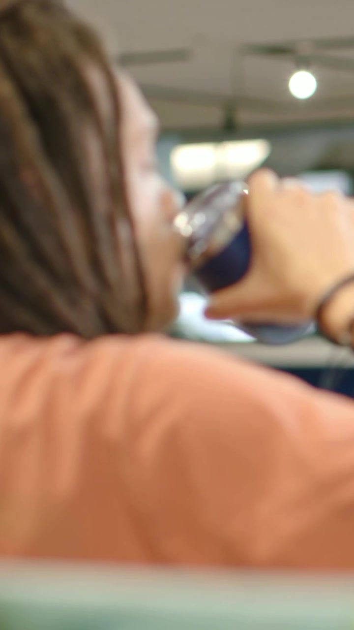 Vertical video: Beginning man lifting blue can and sipping to hydrate at cafe booth, coral tee locs