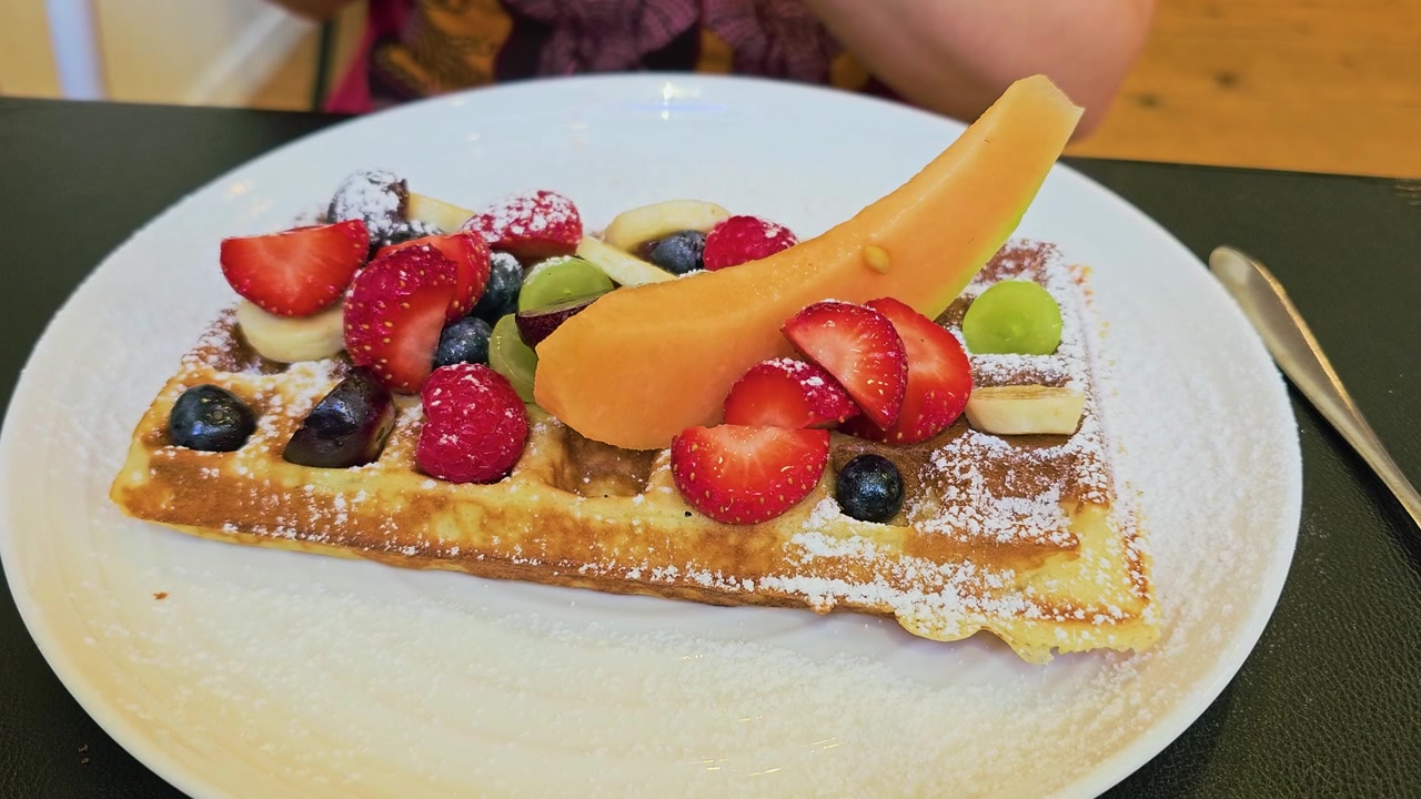 Delizioso waffle per la colazione servito con frutti di bosco colorati, fette di banana, melone, uva e zucchero a velo su un piatto bianco, perfetto per brunch, dessert, cibo da caffè o video di viaggio culinari