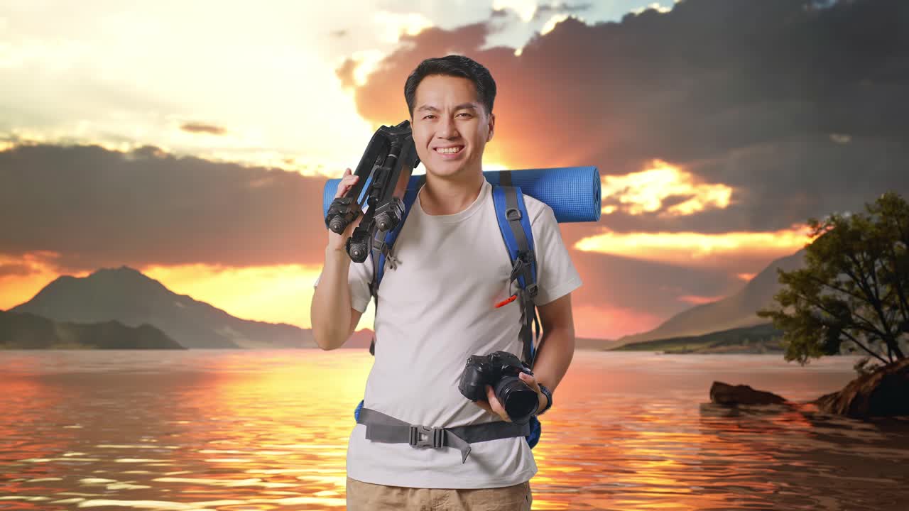 Asian Male Hiker With Mountaineering Backpack Smiling, Holding A Camera, And Putting A Tripod On His Shoulder At A Lake