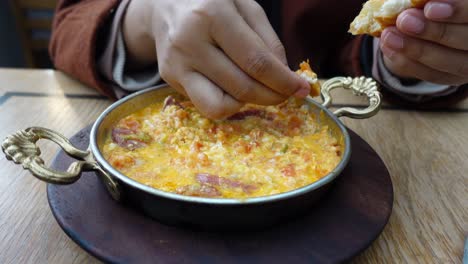 una toma de primer plano de las manos de una persona sosteniendo un pedazo de pan y sumergiéndolo en un cuenco de menemen (un plato turco de huevos, tomates, pimientos y salchichas).