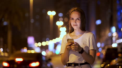 a young woman at night in the city calls a taxi through a smartphone while looking into the screen of a mobile phone and pointing out the coordinates and address of the trip.