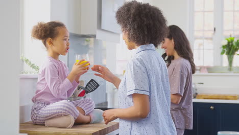 Pregnant-Family-With-Two-Mums-Wearing-Pyjamas-Making-Morning-Pancakes-In-Kitchen-With-Daughter