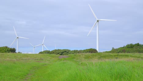 Moving-across-grassy-meadow-towards-rotating-wind-turbines-on-cloudy-summer-day