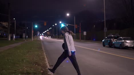 Teenage-girl-in-grey-sweater-with-black-hair-crosses-road-with-basketball-in-hand-at-night