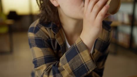 close-up of a girl student with curly hair and glasses drinks coffee from a paper cup while working in the university library