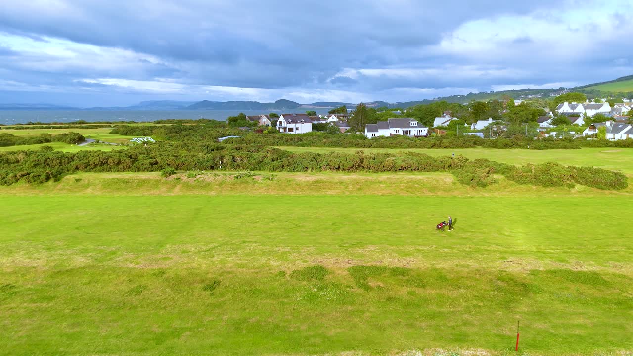 Two golfers and a caddy traverse a vibrant green golf course under dramatic skies, with distant white cottages and rolling hills in Rosemarkie, Scotland